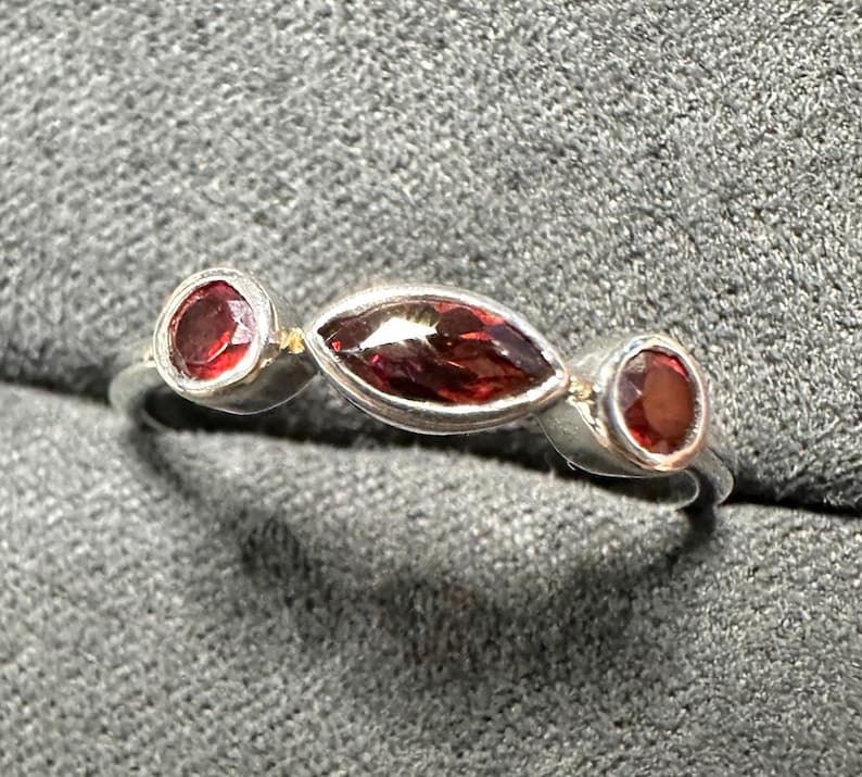 May include: A silver ring featuring three red gemstones. The ring has a central marquise-cut stone flanked by two round stones. The gemstones are set in a silver band, creating a simple yet elegant design.
