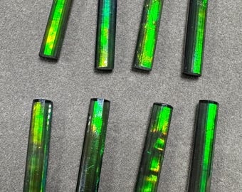 Peut inclure: Huit pierres précieuses rectangulaires et polies, avec une base sombre et des reflets irisés verts et jaunes vibrants. Les pierres sont disposées sur une surface grise, mettant en valeur leur forme cylindrique polie.