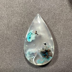 Puede incluir: Un colgante en forma de l&aacute;grima hecho de cuarzo transparente con inclusiones de minerales azules y negros.
