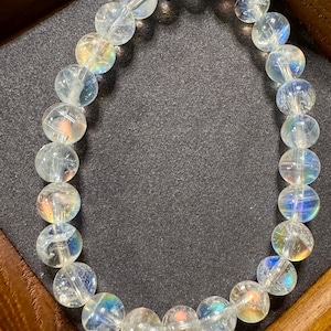 Pode incluir: Uma pulseira de contas de quartzo transparente com flashes iridescentes de arco-&iacute;ris. As contas redondas s&atilde;o enfiadas juntas, criando um acess&oacute;rio delicado e brilhante. A pulseira &eacute; exibida em uma superf&iacute;cie cinza escuro dentro de uma moldura de madeira.