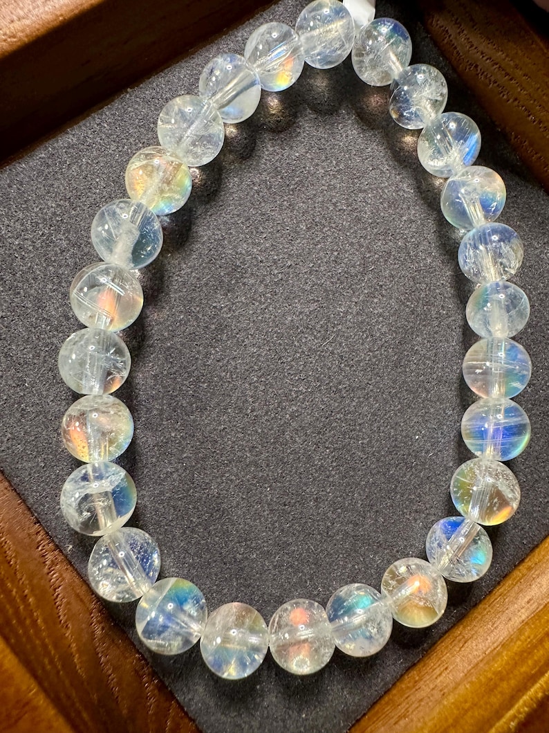 Pode incluir: Uma pulseira de contas de quartzo transparente com flashes iridescentes de arco-&iacute;ris. As contas redondas s&atilde;o enfiadas juntas, criando um acess&oacute;rio delicado e brilhante. A pulseira &eacute; exibida em uma superf&iacute;cie cinza escuro dentro de uma moldura de madeira.