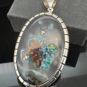 May include: A silver oval pendant with a large, clear, oval-shaped stone. The stone has a cloudy, white background with flecks of copper, green, and blue.