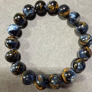 May include: A bracelet made of round, polished beads. The beads are primarily dark blue with swirling patterns of lighter blue and gold flecks. The bracelet is displayed on a gray surface.