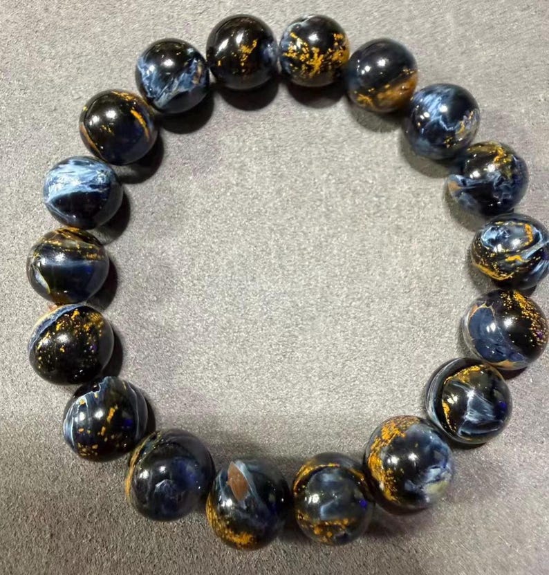 May include: A bracelet made of round, polished beads. The beads are primarily dark blue with swirling patterns of lighter blue and gold flecks. The bracelet is displayed on a gray surface.