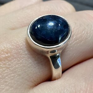 May include: A silver ring featuring a large, oval-shaped, dark blue gemstone. The gemstone is set in a simple, polished silver band. The ring is shown on a person's finger, highlighting the design and color of the jewelry.