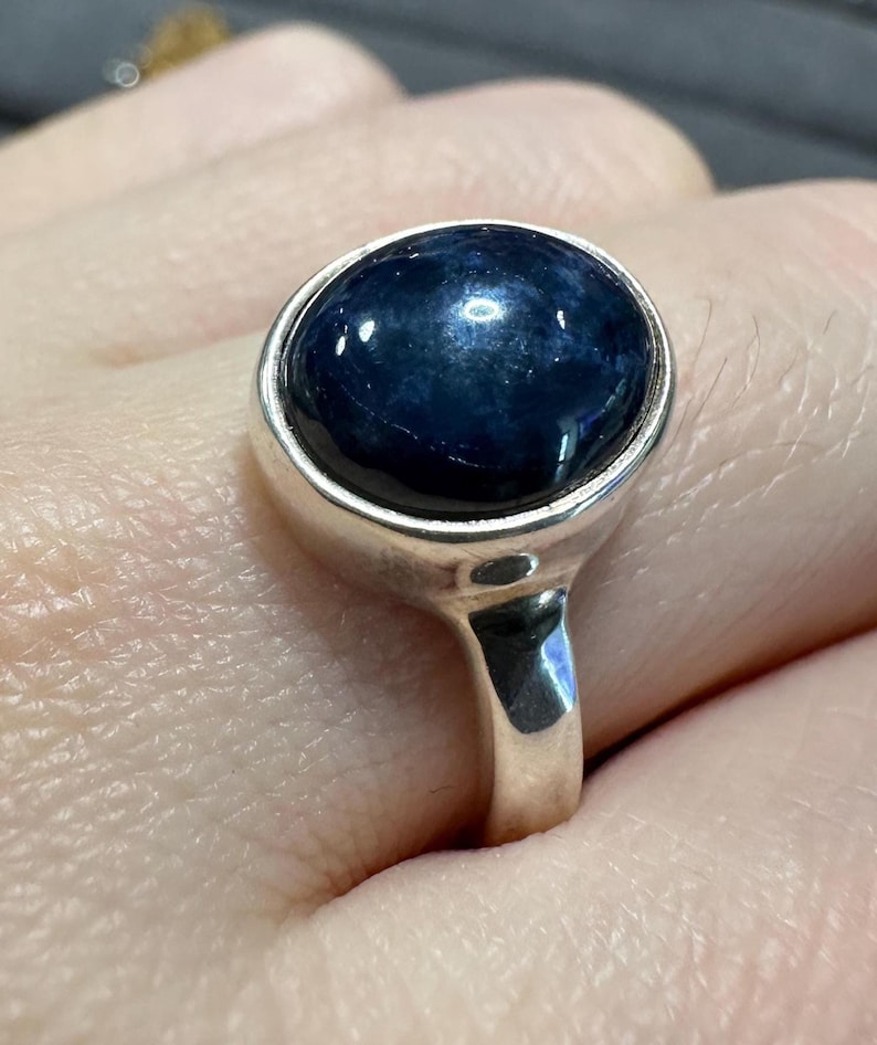 May include: A silver ring featuring a large, oval-shaped, dark blue gemstone. The gemstone is set in a simple, polished silver band. The ring is shown on a person's finger, highlighting the design and color of the jewelry.