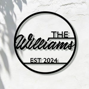 Puede incluir: Letrero de nombre familiar de metal negro con el nombre "Williams" en fuente cursiva, encima de "THE" y debajo de "EST 2024" dentro de un círculo negro. El letrero está montado en una pared blanca texturizada, con follaje verde en el fondo.