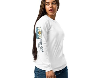 May include: White long-sleeve shirt with a blue and yellow graphic on the left sleeve. The graphic features a sun, waves, and the text "SUNNYSIDE COTTAGES OCONTO, WI".
