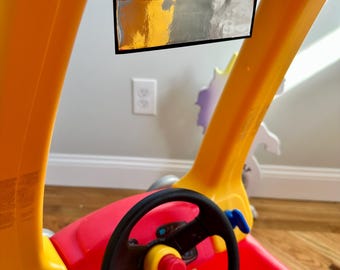 May include: A red and yellow toy car with a black steering wheel and a blue and silver rearview mirror. The car has a yellow dashboard with a black steering wheel and a blue and silver rearview mirror.