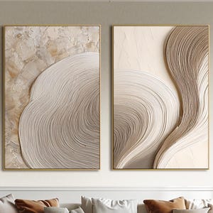 Puede incluir: Dos obras de arte murales abstractas enmarcadas. La obra presenta patrones texturizados y arremolinados en tonos beige y crema neutros, sobre un marco dorado. El arte se exhibe sobre un sofá grande y cómodo con varios cojines.
