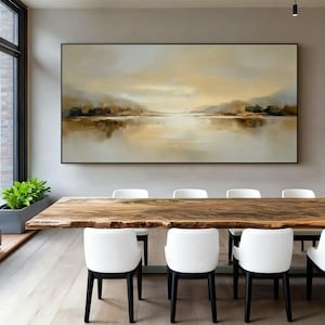 May include: An abstract landscape painting in a black frame, depicting a serene lake scene with muted tones of beige, brown, and gold. The artwork hangs above a large wooden dining table with white chairs.