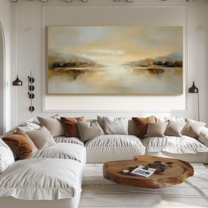 May include: A large landscape painting in a gold frame depicts a serene waterscape with muted tones of beige, brown, and gray. The artwork hangs above a white sectional sofa adorned with various throw pillows in neutral colors. A wooden coffee table sits in front of the sofa.