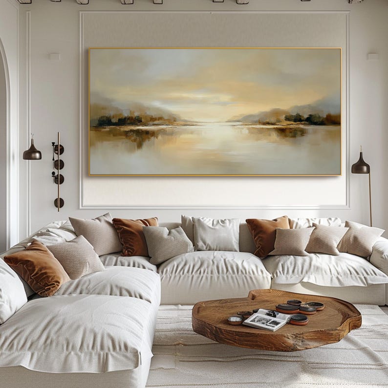 May include: A large landscape painting in a gold frame depicts a serene waterscape with muted tones of beige, brown, and gray. The artwork hangs above a white sectional sofa adorned with various throw pillows in neutral colors. A wooden coffee table sits in front of the sofa.