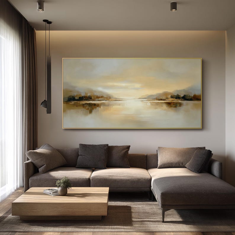 May include: A large rectangular landscape painting with a gold frame. The artwork depicts a serene lake scene with muted tones of beige, brown, and gold. The painting hangs above a brown sectional sofa and a wooden coffee table.