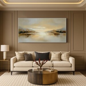 May include: A landscape painting with a serene lake scene in shades of gold, brown, and beige hangs above a cream-colored sofa. The room features a round coffee table, decorative pillows, and a table lamp, creating a warm and inviting atmosphere.
