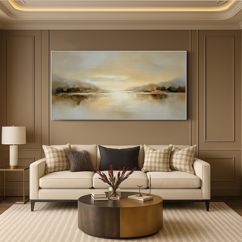 May include: A landscape painting with a serene lake scene in shades of gold, brown, and beige hangs above a cream-colored sofa. The room features a round coffee table, decorative pillows, and a table lamp, creating a warm and inviting atmosphere.