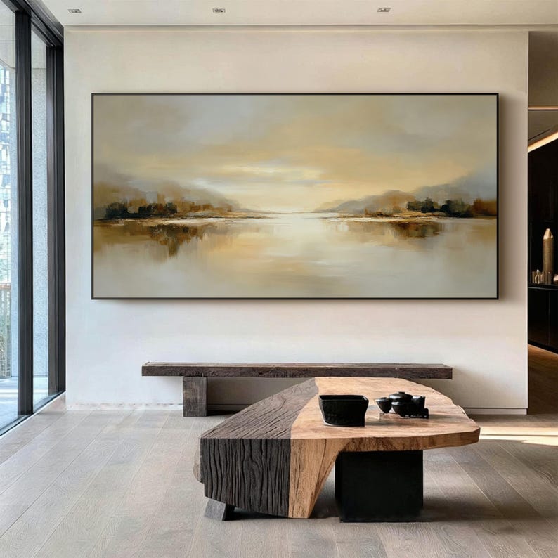 May include: Large rectangular landscape painting with a black frame. The artwork depicts a serene lake scene with muted tones of beige, brown, and gold. The water reflects the sky and distant hills, creating a sense of calm.