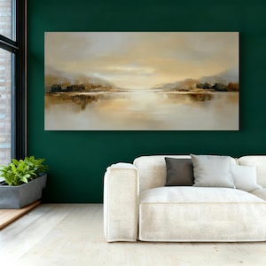 May include: An abstract landscape painting with a serene lake scene. The artwork features muted tones of beige, brown, and gold, with reflections in the water. The painting is displayed on a dark green wall above a white sofa.