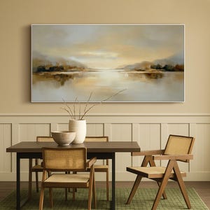 May include: A large landscape painting in a white frame depicts a serene lake scene with muted tones of beige, brown, and gray. The artwork hangs above a dark wood dining table with four chairs, featuring woven cane backs and seats. A white vase and a wooden bowl are on the table.