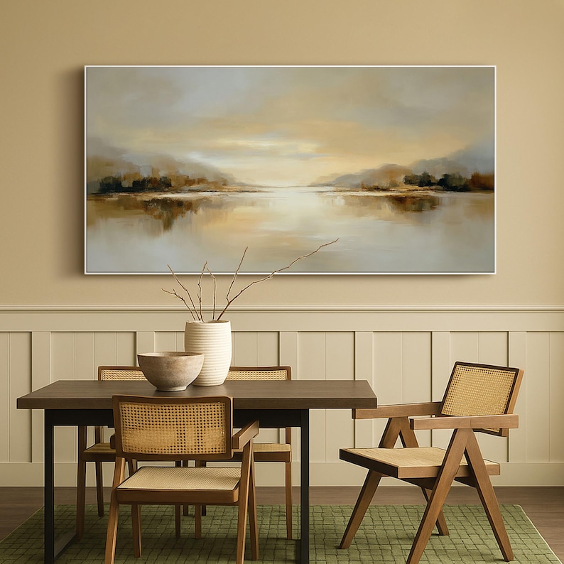 May include: A large landscape painting in a white frame depicts a serene lake scene with muted tones of beige, brown, and gray. The artwork hangs above a dark wood dining table with four chairs, featuring woven cane backs and seats. A white vase and a wooden bowl are on the table.