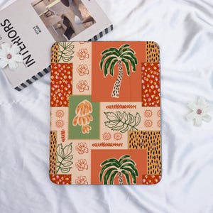 May include: An orange, green, and beige tablet case with a patchwork design. The design includes palm trees, flowers, and leaf patterns. The case is on a white surface with a book and a flower in the background.