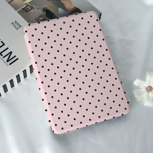 Könnte beinhalten: Eine hellrosa Tablet-Hülle mit einem sich wiederholenden Muster aus kleinen, dunkelbraunen Polka-Dots. Die rechteckige Hülle hat abgerundete Ecken. Die Hülle liegt auf einer weißen Oberfläche neben einem Buch mit dem Wort "STYLE BOOK" auf dem Buchrücken.