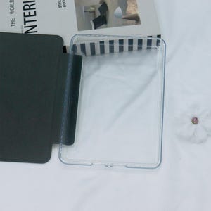 Może przedstawiać: Przezroczyste plastikowe etui na tablet lub czytnik e-book&oacute;w. Etui ma czarną klapkę, kt&oacute;ra składa się na przednią część urządzenia.