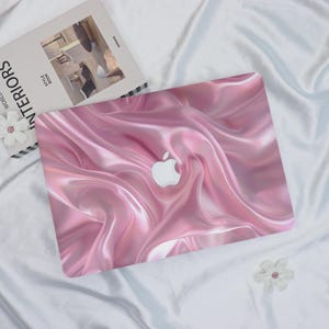 May include: A pink laptop cover with a glossy, flowing fabric design. The cover features the Apple logo in white. The design resembles draped silk or satin, creating a luxurious aesthetic. A book and a flower are in the background.