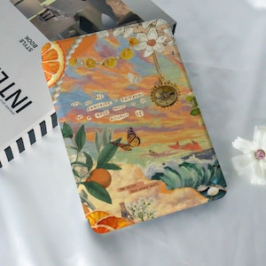 May include: A colorful collage style notebook cover with a butterfly, oranges, and a compass. The text on the cover reads "It's not all sunshine & rainbows but a good amount of it actually is." and "Be wild. But stay soft."