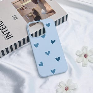 May include: A light blue phone case with a pattern of dark blue hearts. The case is lying on a white surface next to a book and white flowers.