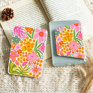 May include: Two rectangular card cases featuring a vibrant floral pattern with pink, yellow, orange, and green flowers on a white background. The cases are displayed on an open book with text and a small pinecone, resting on a textured lace fabric.