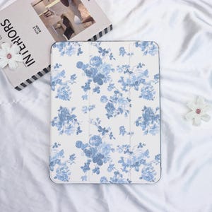 May include: A white tablet case with a blue floral pattern. The design features clusters of blue roses and leaves on a white background. The case is rectangular and has a black border.
