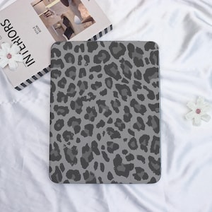 Könnte beinhalten: Eine graue Tablet-Hülle mit Leopardenmuster. Die Hülle hat ein Muster aus dunkelgrauen Flecken auf hellgrauem Hintergrund. Die Hülle ist rechteckig mit abgerundeten Ecken. Das Bild enthält auch ein Buch und eine Blume.