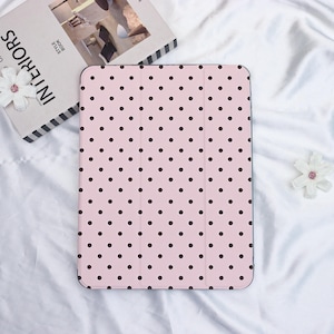 May include: Pink tablet case featuring a pattern of dark brown polka dots. The case is open, showing the inside. It rests on a white surface, beside a book and a flower. The case is designed to protect a tablet.