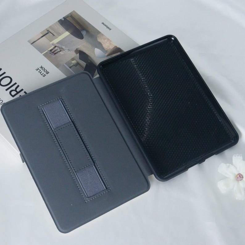 May include: A grey, textured, protective case for an e-reader. The case has a built-in stand and a strap to hold the e-reader in place. The case is open, revealing a black, textured interior.