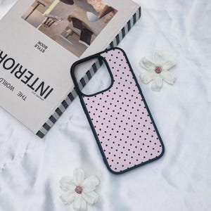 May include: A phone case with a light pink background and a pattern of small black polka dots. The case has a black border and is next to a book titled "INTERIOR" and two white flowers.