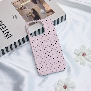 May include: A pale pink phone case featuring a polka dot design in black. The case is displayed on a white surface, alongside a book and white flowers.