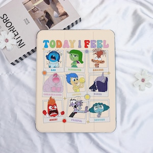 May include: A beige tablet case with the text "TODAY I FEEL" in bright colours. The case shows cartoon characters representing emotions such as sad, disgusted, anxious, and joyful. The case is on a white surface.