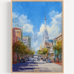 Op de afbeelding: Aquarel schilderij van een stadsstraatbeeld met een hoge kerktoren op de achtergrond. Het kunstwerk toont een blauwe lucht met pluizige witte wolken, gebouwen, bomen en auto's op de weg. De schildering is ingelijst in een lichte houten lijst.