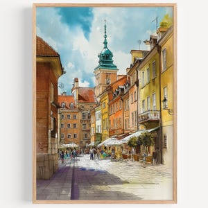 May include: Watercolor print of a European city street scene. The artwork features colorful buildings, a tall tower, and people walking along a cobblestone street. The sky is blue with fluffy clouds, and the print is framed in a light wood frame.
