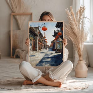 May include: A framed watercolor print depicting a traditional Asian street scene. The artwork features buildings with dark roofs, red lanterns, and a cobblestone street. The print is held by a person sitting cross-legged.