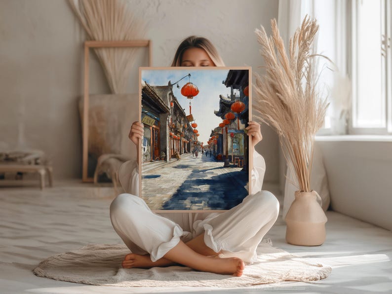 May include: A framed watercolor print depicting a traditional Asian street scene. The artwork features buildings with dark roofs, red lanterns, and a cobblestone street. The print is held by a person sitting cross-legged.