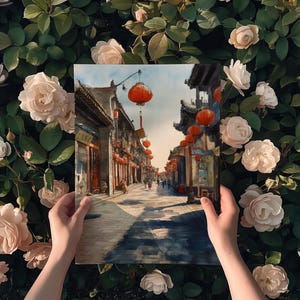 May include: A painting depicting a street scene with traditional Chinese architecture, red lanterns, and figures. The artwork is held against a backdrop of lush green foliage and soft pink roses.