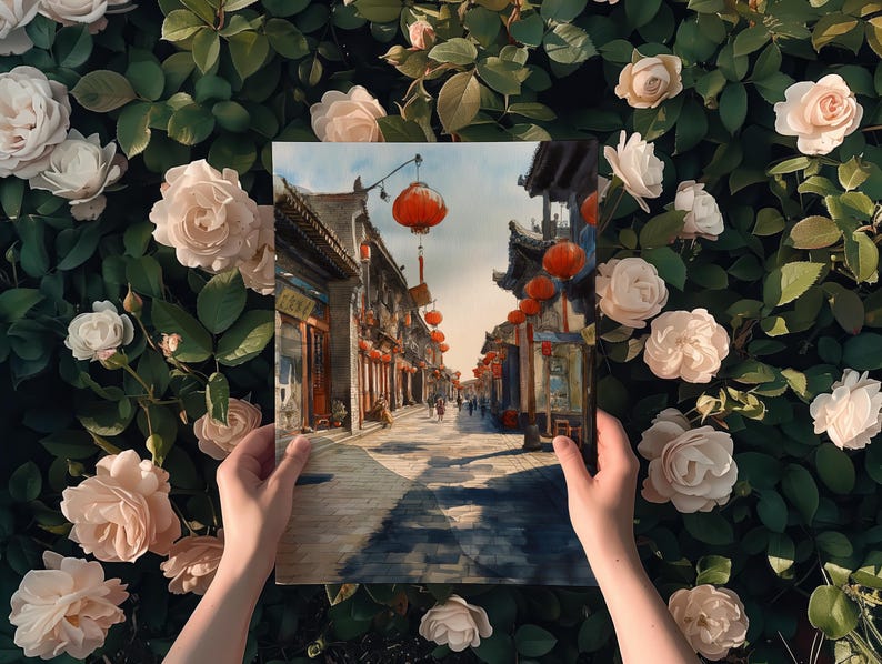May include: A painting depicting a street scene with traditional Chinese architecture, red lanterns, and figures. The artwork is held against a backdrop of lush green foliage and soft pink roses.