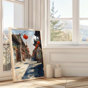 May include: A framed watercolor print of a traditional Asian street scene. The artwork depicts buildings with ornate roofs, red lanterns, and a cobblestone street. The print is leaning against a window, with two white vases in front.