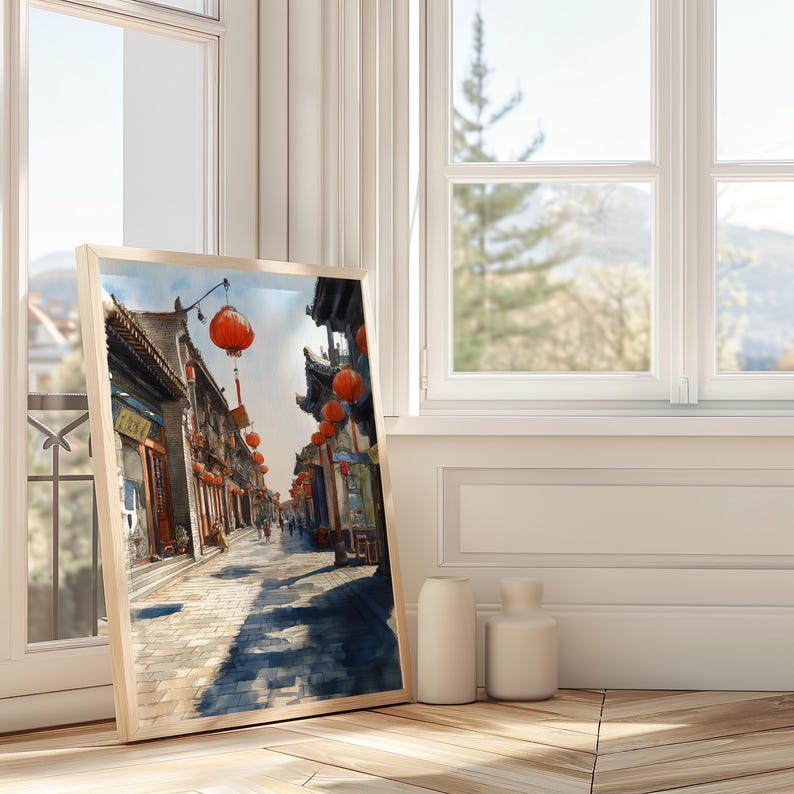 May include: A framed watercolor print of a traditional Asian street scene. The artwork depicts buildings with ornate roofs, red lanterns, and a cobblestone street. The print is leaning against a window, with two white vases in front.