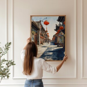 May include: A framed watercolor painting of a traditional Asian street scene with red lanterns hanging overhead. The artwork depicts buildings, shops, and people on a sunny day. The painting is being held up against a white wall.