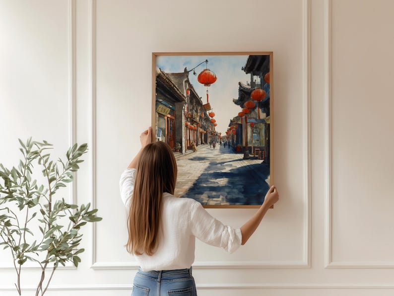 May include: A framed watercolor painting of a traditional Asian street scene with red lanterns hanging overhead. The artwork depicts buildings, shops, and people on a sunny day. The painting is being held up against a white wall.