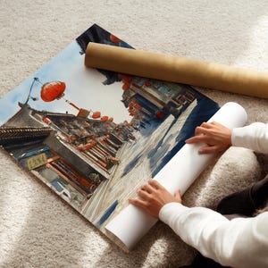 May include: A watercolor print of a traditional Asian street scene with red lanterns and detailed architecture. The print is partially rolled, with a brown tube and a white tube nearby. The scene is set against a blue sky.