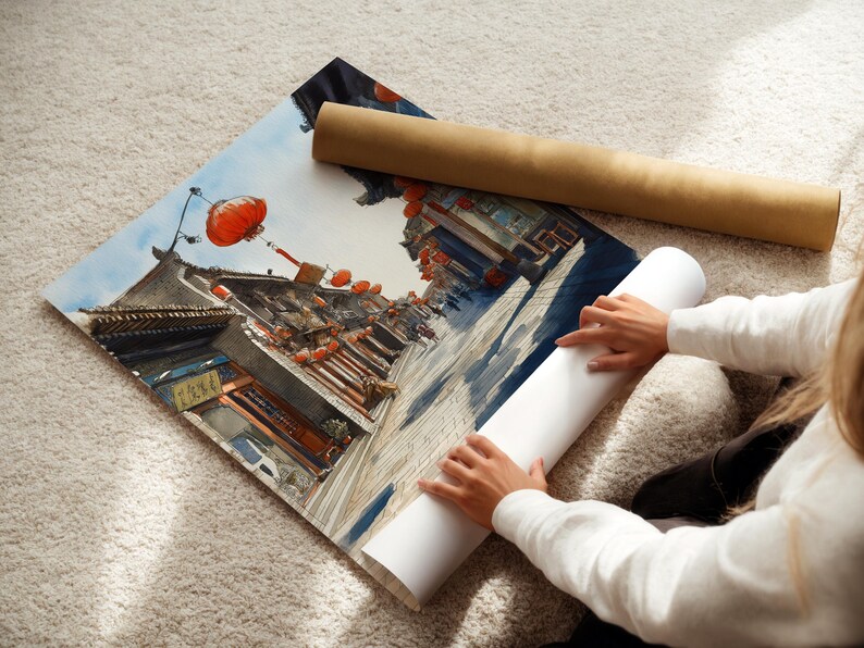 May include: A watercolor print of a traditional Asian street scene with red lanterns and detailed architecture. The print is partially rolled, with a brown tube and a white tube nearby. The scene is set against a blue sky.